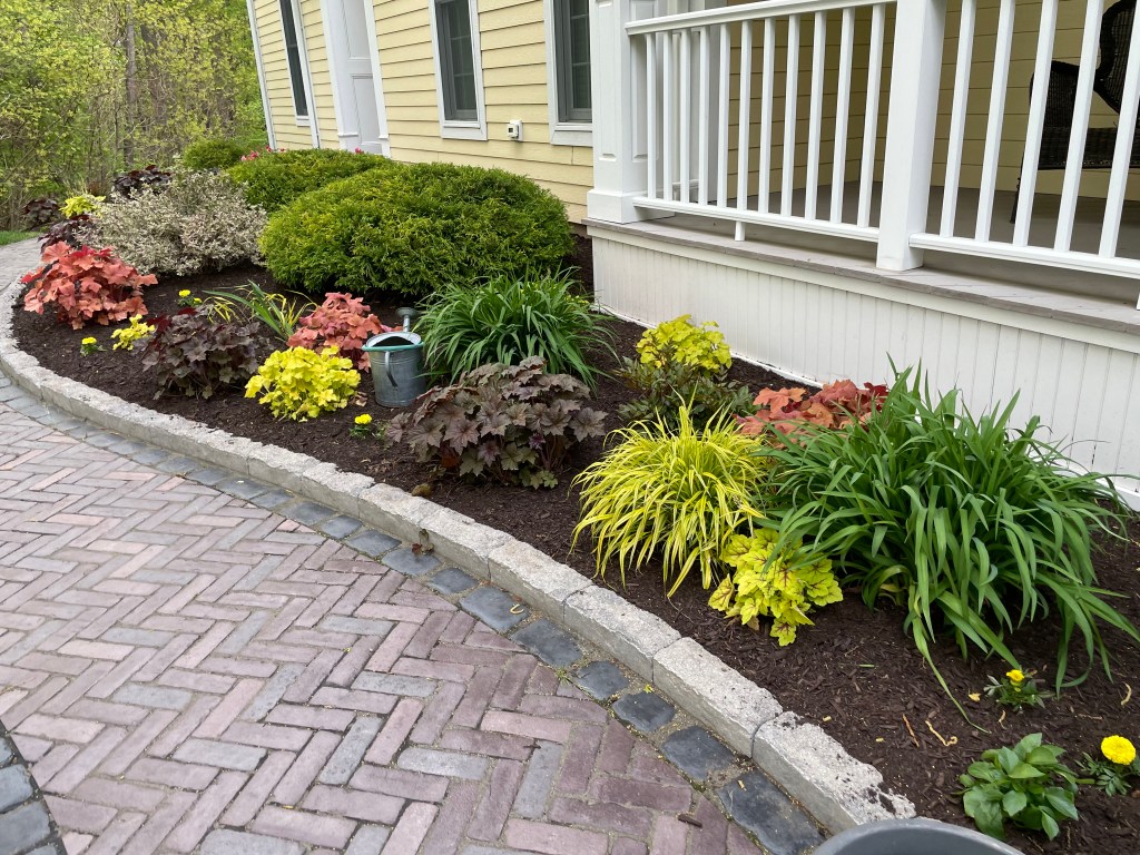 Landscape project | Larson’s Garden Center and Landscaping A colorful flowerbed featuring a variety of plants, including green bushes, colorful foliage, and yellow flowers, alongside a brick pathway.